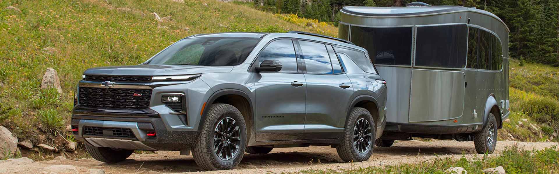 A Chevrolet Traverse SUV towing a sleek, modern Airstream travel trailer along a rugged, grassy terrain. The vehicle is equipped with off-road tires and has a bold, athletic design, while the trailer is compact and stylish, featuring a shiny metallic exterior.